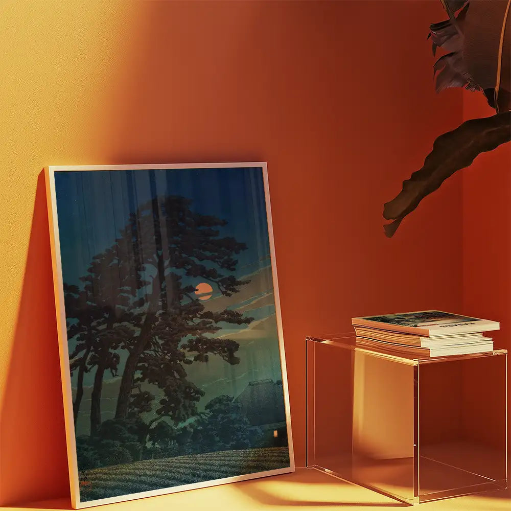 Framed painting of a nighttime landscape featuring trees and a setting sun, displayed against an orange wall with a stack of books in a clear acrylic holder.