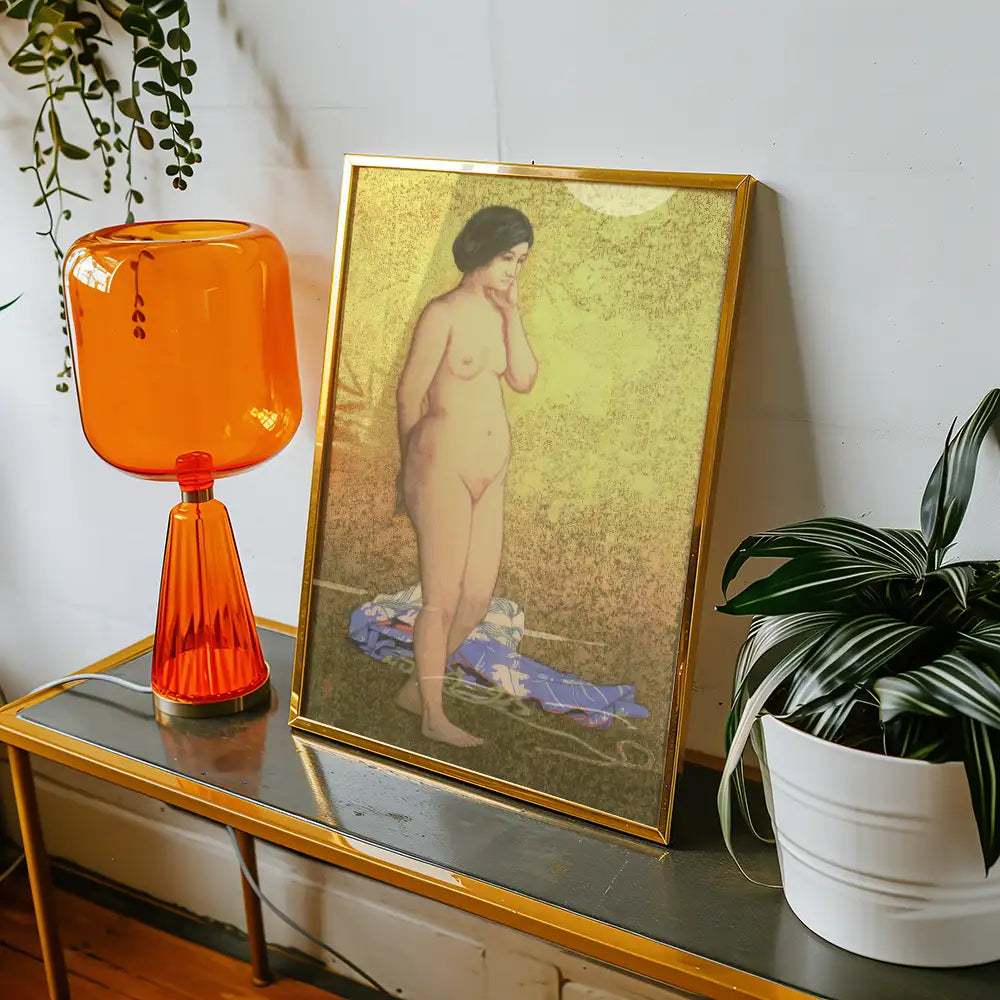 Nude painting of a woman framed in gold, displayed on a stylish table with an orange lamp and a potted plant.