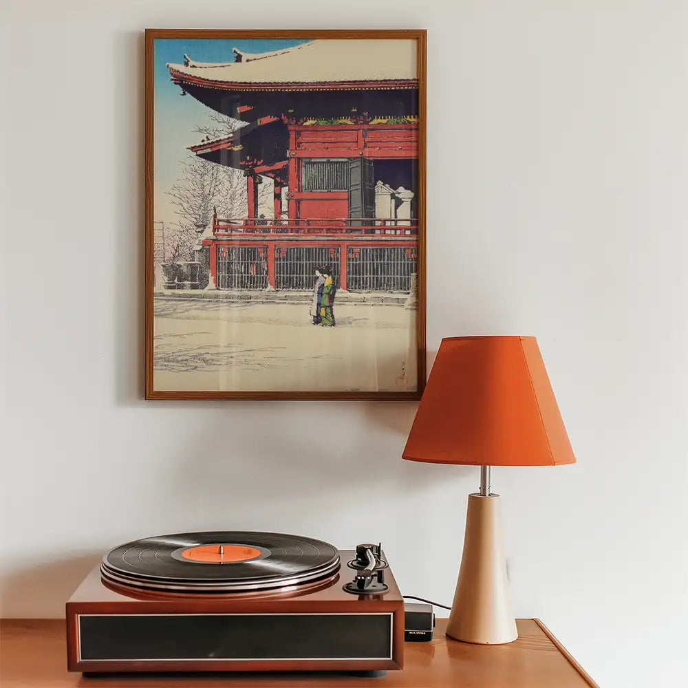 Framed painting of a traditional building with two figures in front, displayed above a record player and an orange lamp.