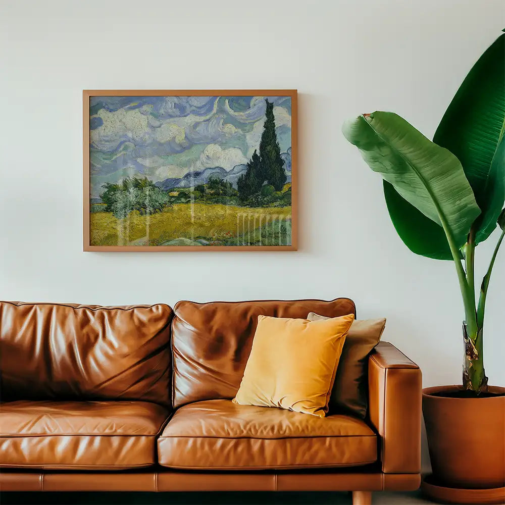 Framed painting of a landscape featuring rolling hills, cypress trees, and a cloudy sky, displayed above a brown leather couch with a yellow accent pillow and a large green plant.