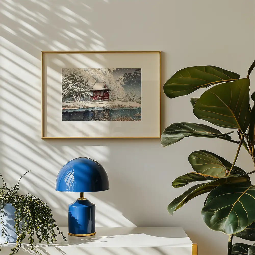 Framed winter landscape painting of a red house by a lake, displayed on a light-colored wall next to a blue lamp and green plants.
