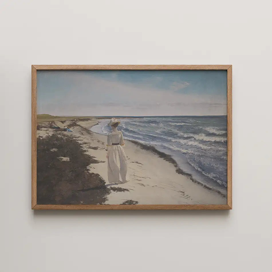 A woman in a white dress and hat walking along a sandy beach with waves crashing on the shore, framed in a wooden frame.