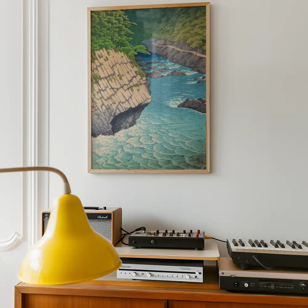 Painting of a serene water scene with rocky cliffs and lush greenery, framed and displayed on a wall above a vintage audio setup and a yellow lamp.