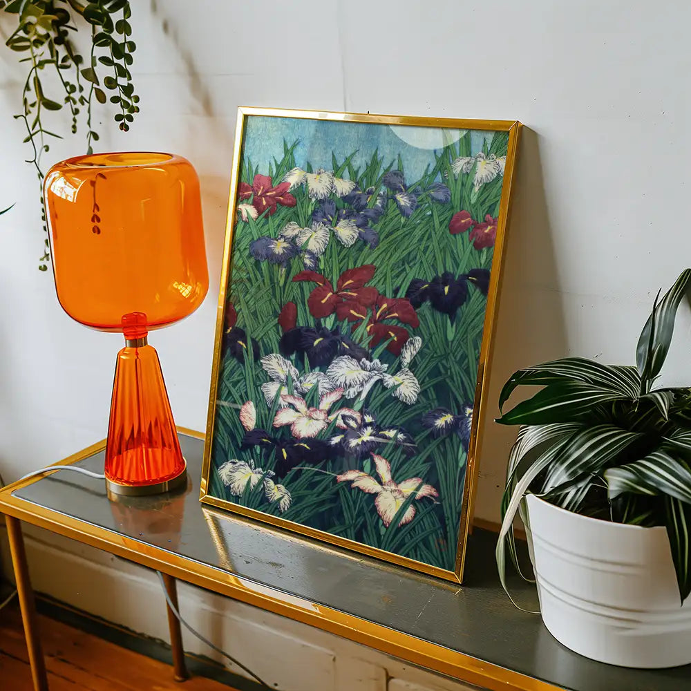 A vibrant framed painting of colorful irises displayed on a shelf next to an orange lamp and a potted plant.