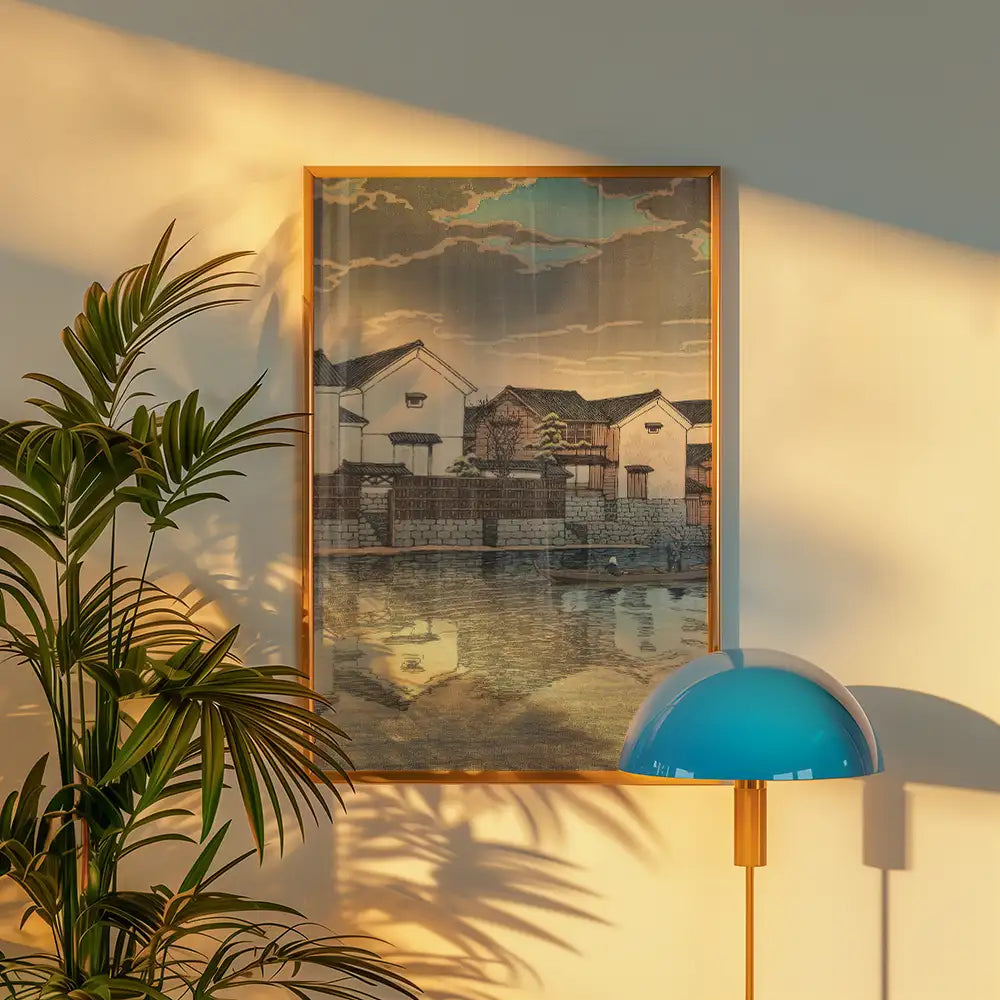 A framed painting of traditional houses beside a calm waterway, illuminated by warm sunlight, accompanied by a potted plant and a stylish blue lamp.