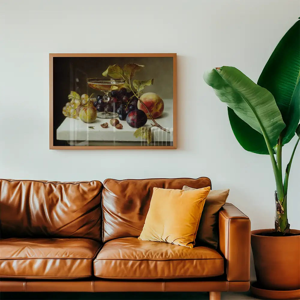 Still life painting of fruit displayed in a glass bowl, framed and hung on a wall above a brown leather sofa, with a potted plant in the foreground.