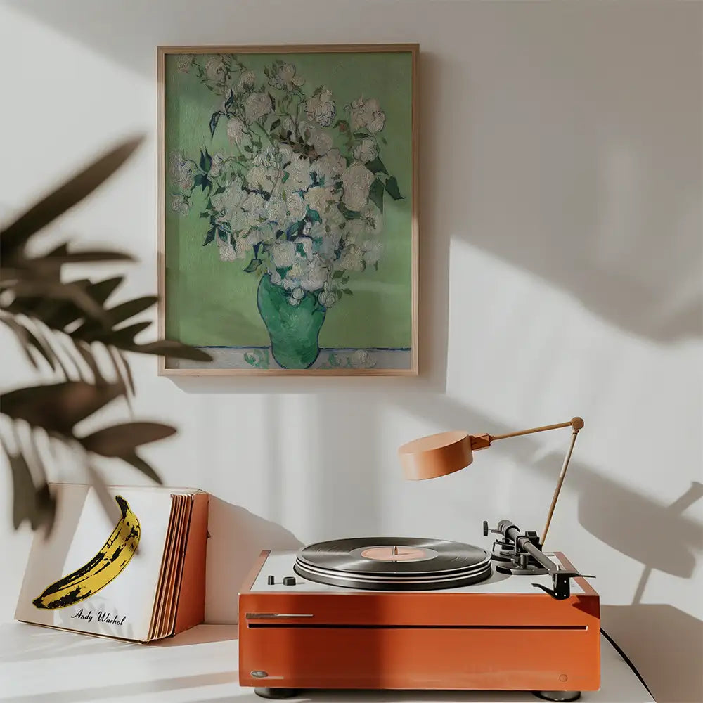 Painting of white flowers in a green vase framed on a wall above a stylish orange record player with a lamp and vinyl records.