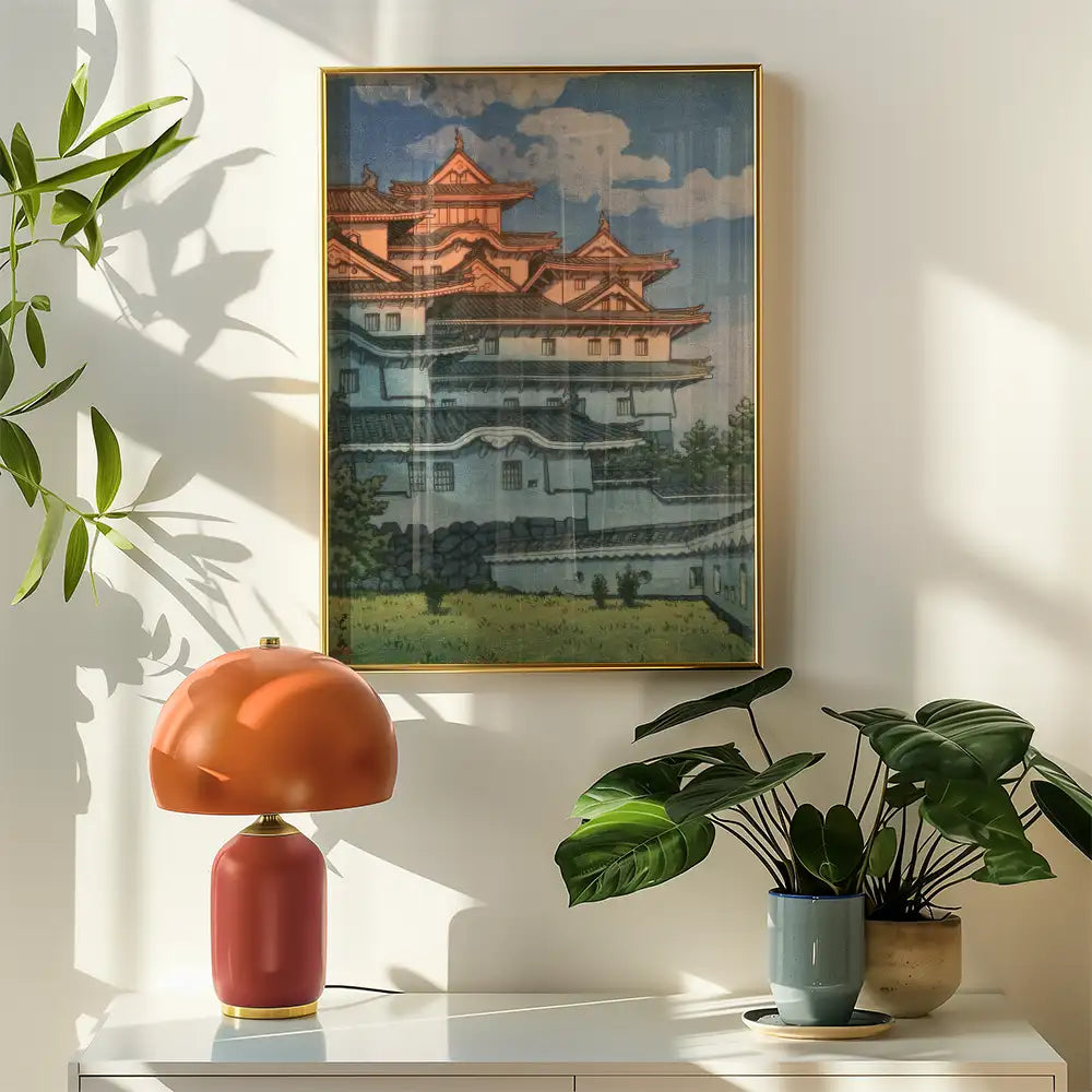 A framed painting of a traditional Japanese castle displayed on a wall next to a modern orange lamp and a green potted plant.