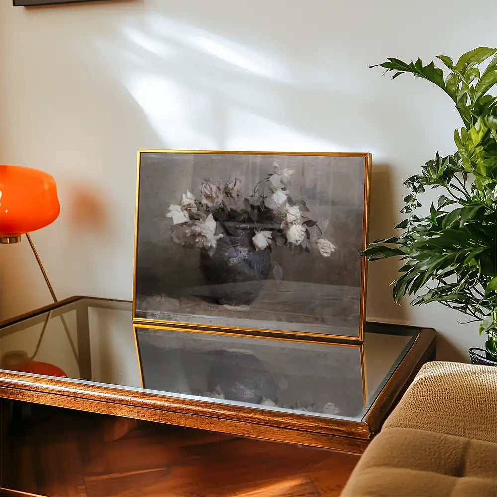 Framed painting of white flowers in a dark vase displayed on a wooden table with a reflective surface, alongside a decorative orange lamp and green plant.