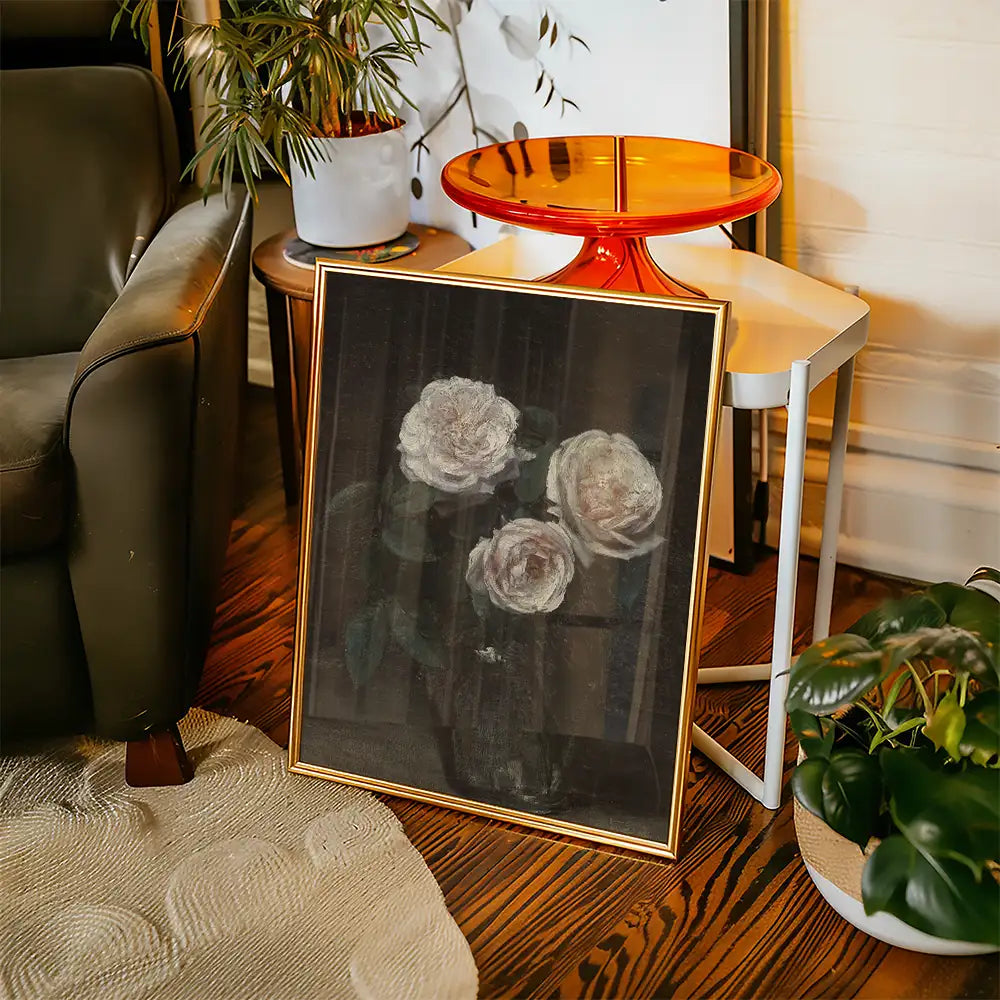 A framed painting of three roses, displayed in a cozy living room setting with plants and furniture.