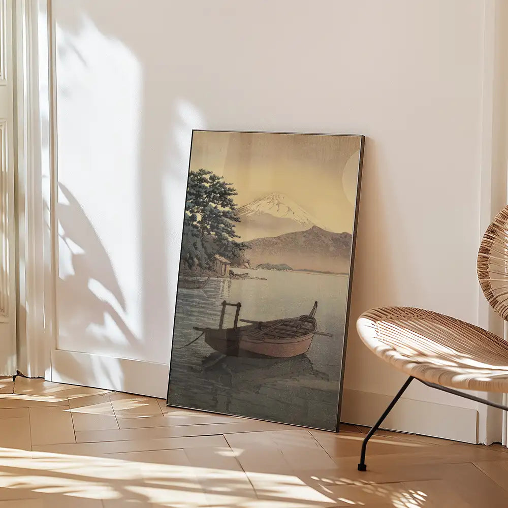 Framed painting of a tranquil landscape featuring a boat on a calm lake with a mountain in the background, leaning against a wall in a bright interior setting.