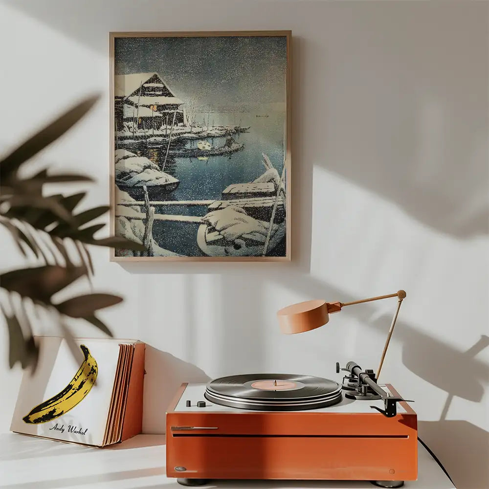 Framed painting of a snowy landscape with boats and a house, displayed above a vintage record player with a lamp and a banana-themed album cover.