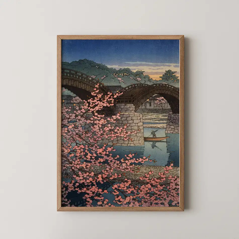 Framed painting of a serene landscape featuring a stone bridge, cherry blossom trees in bloom, and a figure in a boat on a reflective body of water at sunset.