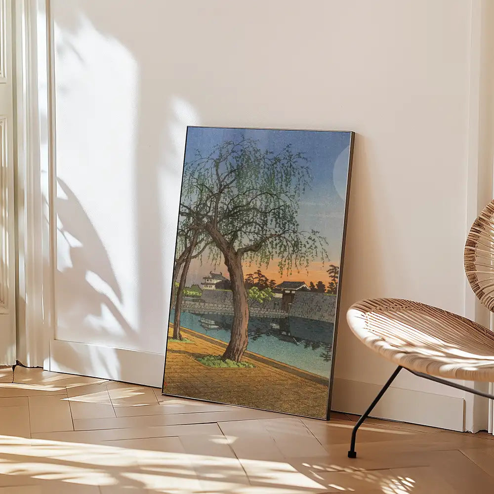 A framed painting of a serene landscape featuring a tree by a river during sunset, leaning against a wall in a stylish interior setting.
