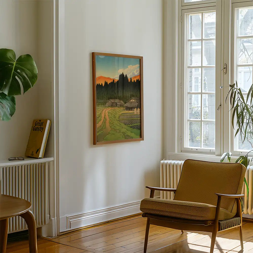 Framed painting of a scenic landscape featuring rolling hills, a dirt path, and rustic cottages, displayed on a wall in a cozy living room with a plant and a chair.