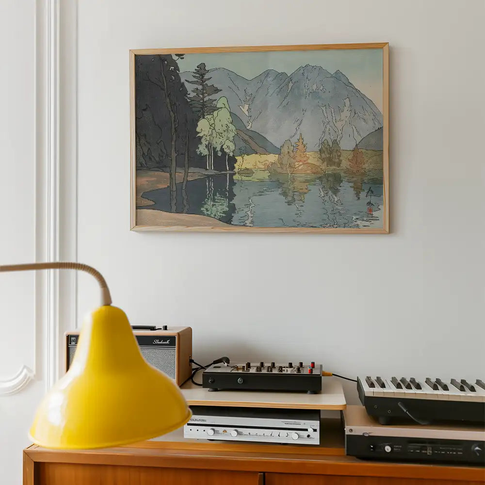 Framed painting of a serene landscape with mountains and trees reflected in a calm lake, hung on a wall above a wooden console table with audio equipment and a yellow lamp.