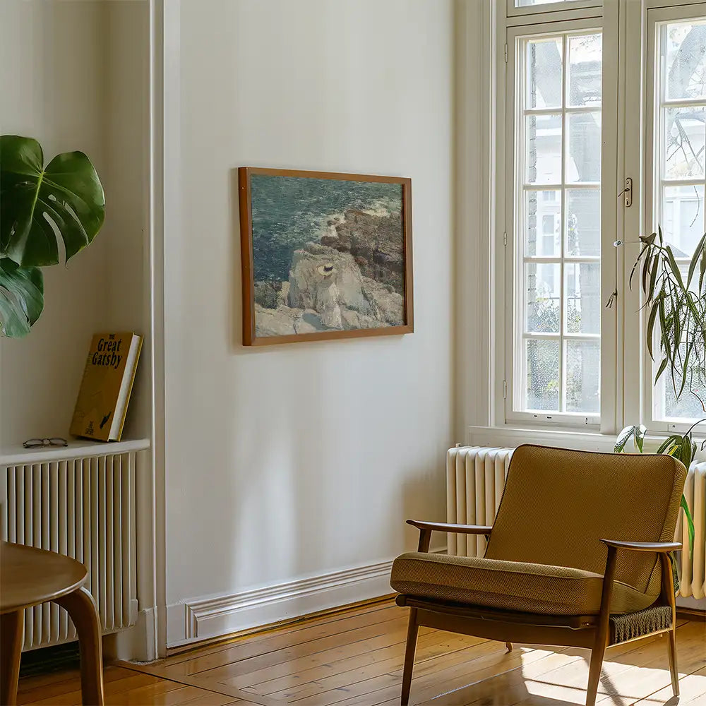 Painting of a rocky shoreline with water, framed and displayed on a wall in a bright room featuring a chair and a potted plant.