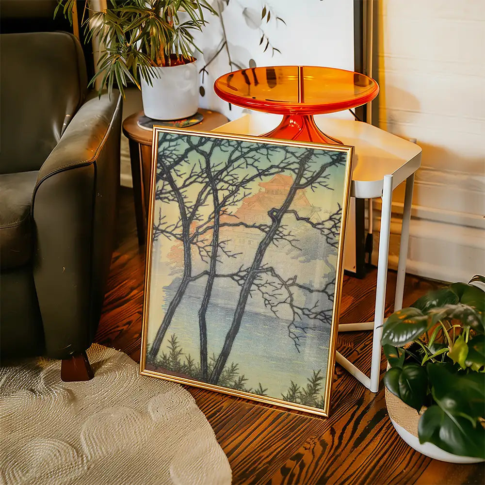 Framed painting of bare trees against a serene sunset backdrop, displayed in a cozy living room setting with a small table and potted plants.