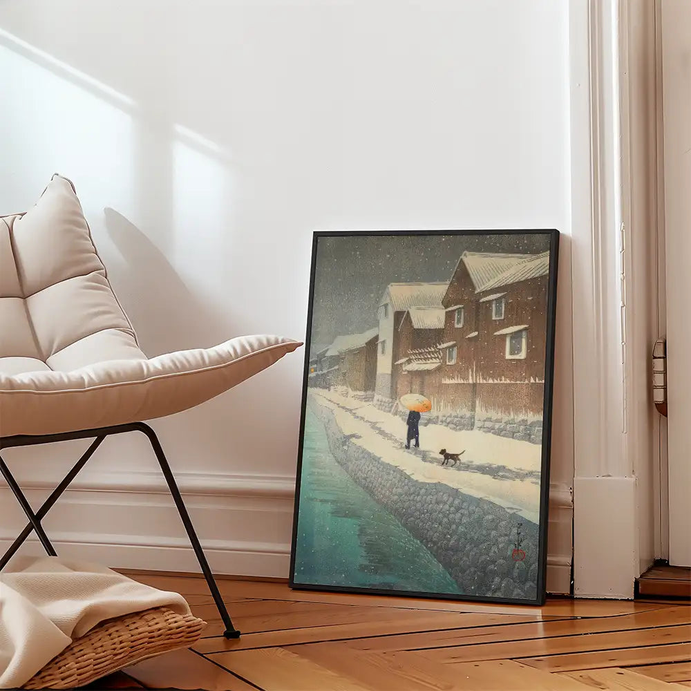 A framed painting of a snowy street scene with a person holding an umbrella and walking a dog, placed next to a beige chair in a modern interior setting.