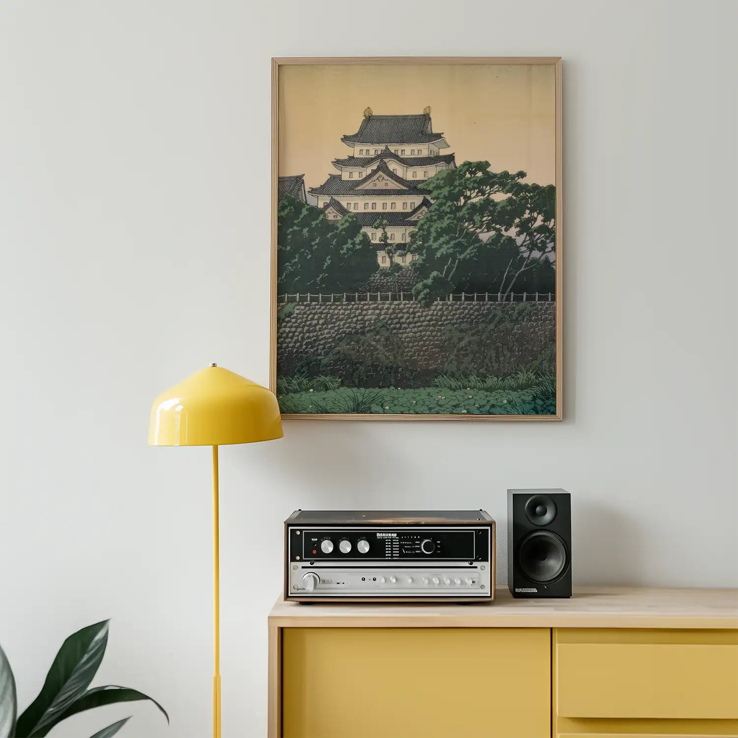 Framed painting of a traditional building surrounded by greenery, displayed on a light wall above a yellow lamp and audio equipment on a wooden console.