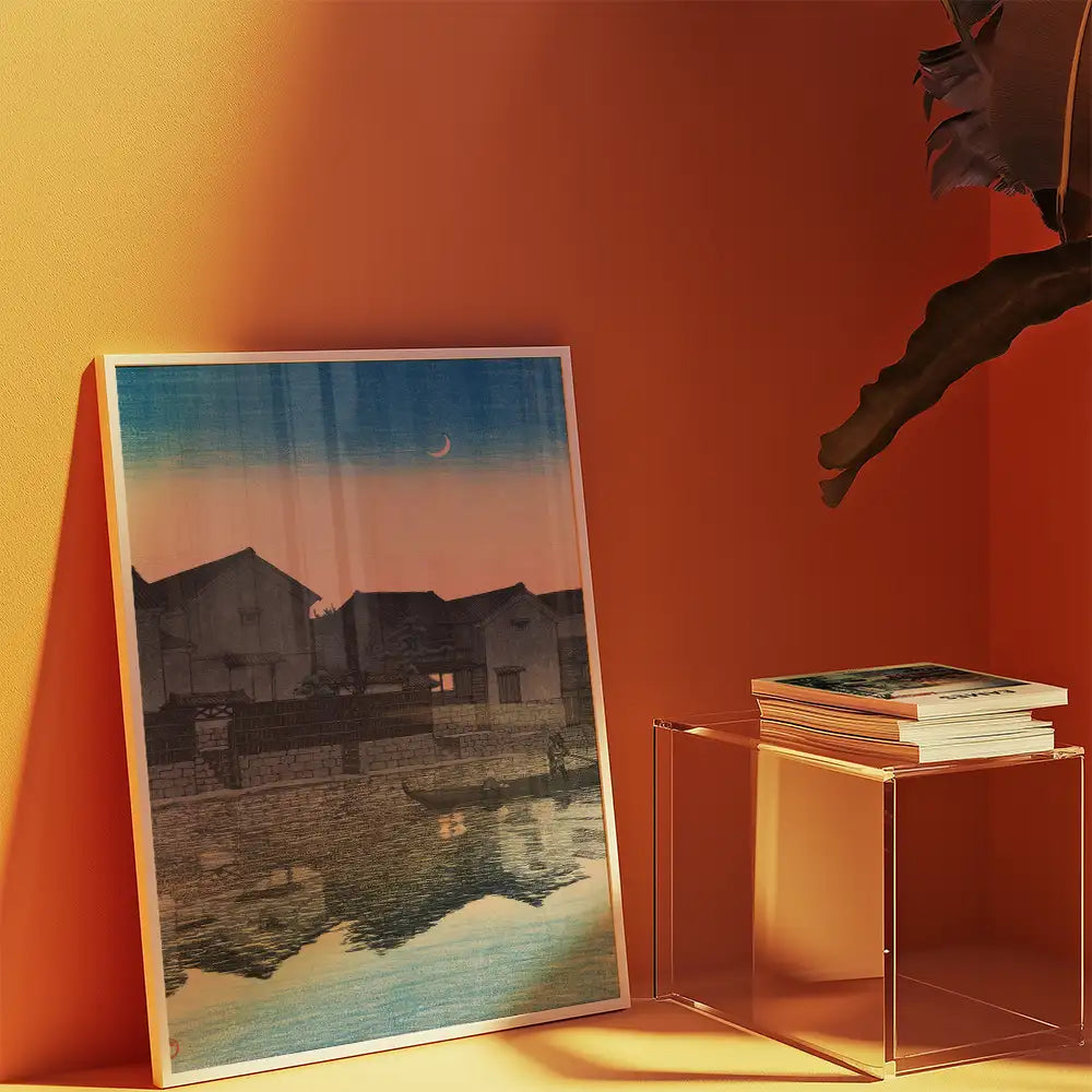 A framed painting of a serene landscape with houses and a crescent moon reflected in the water, leaning against a vibrant orange wall next to a clear acrylic table stacked with books.