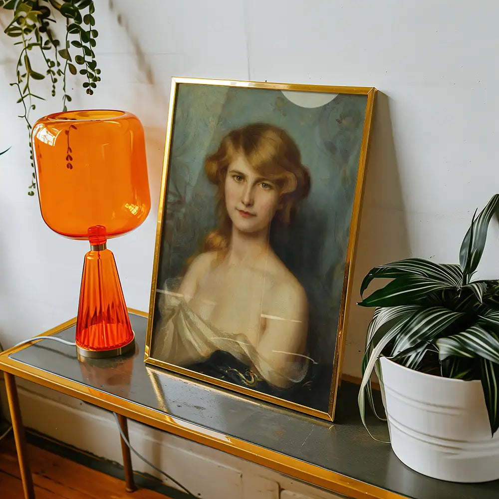 Portrait of a woman in a gold frame placed on a table next to an orange lamp and a potted plant.