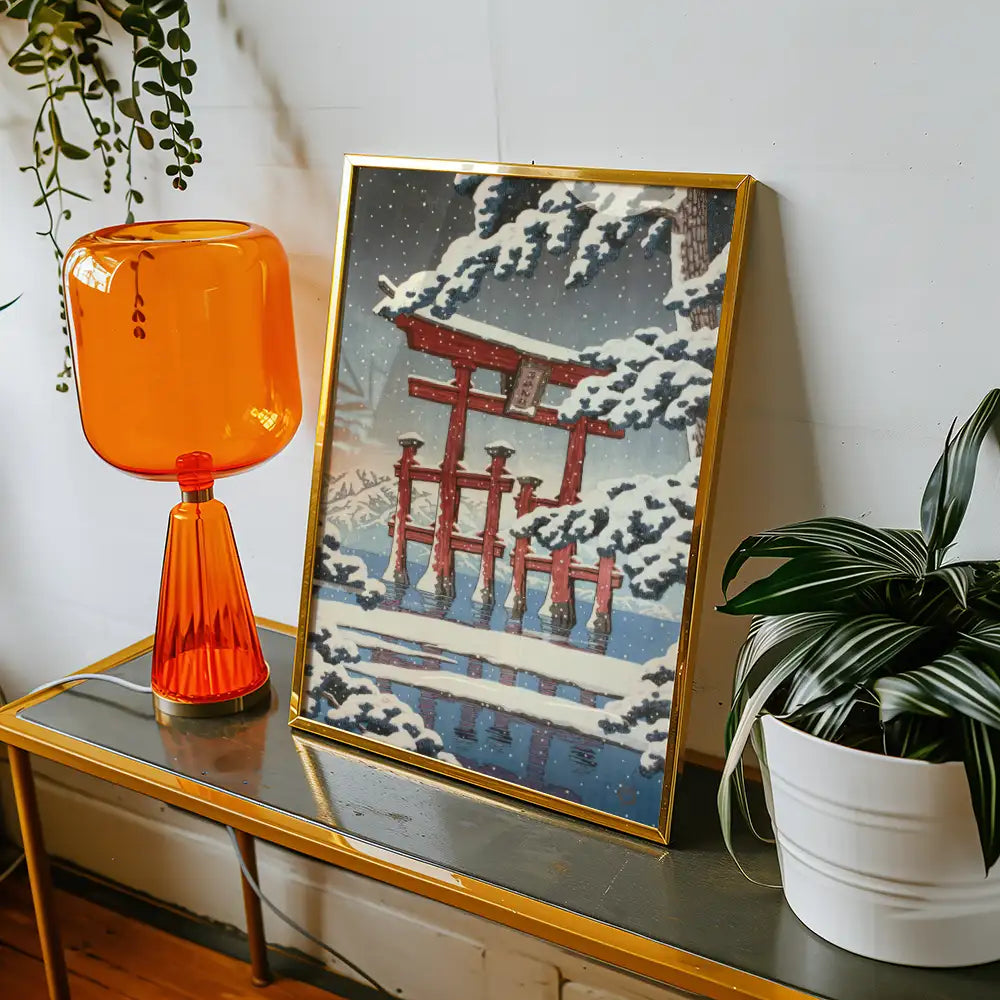 Framed painting of a snowy landscape featuring a traditional red torii gate, displayed on a table alongside an orange lamp and a potted plant.