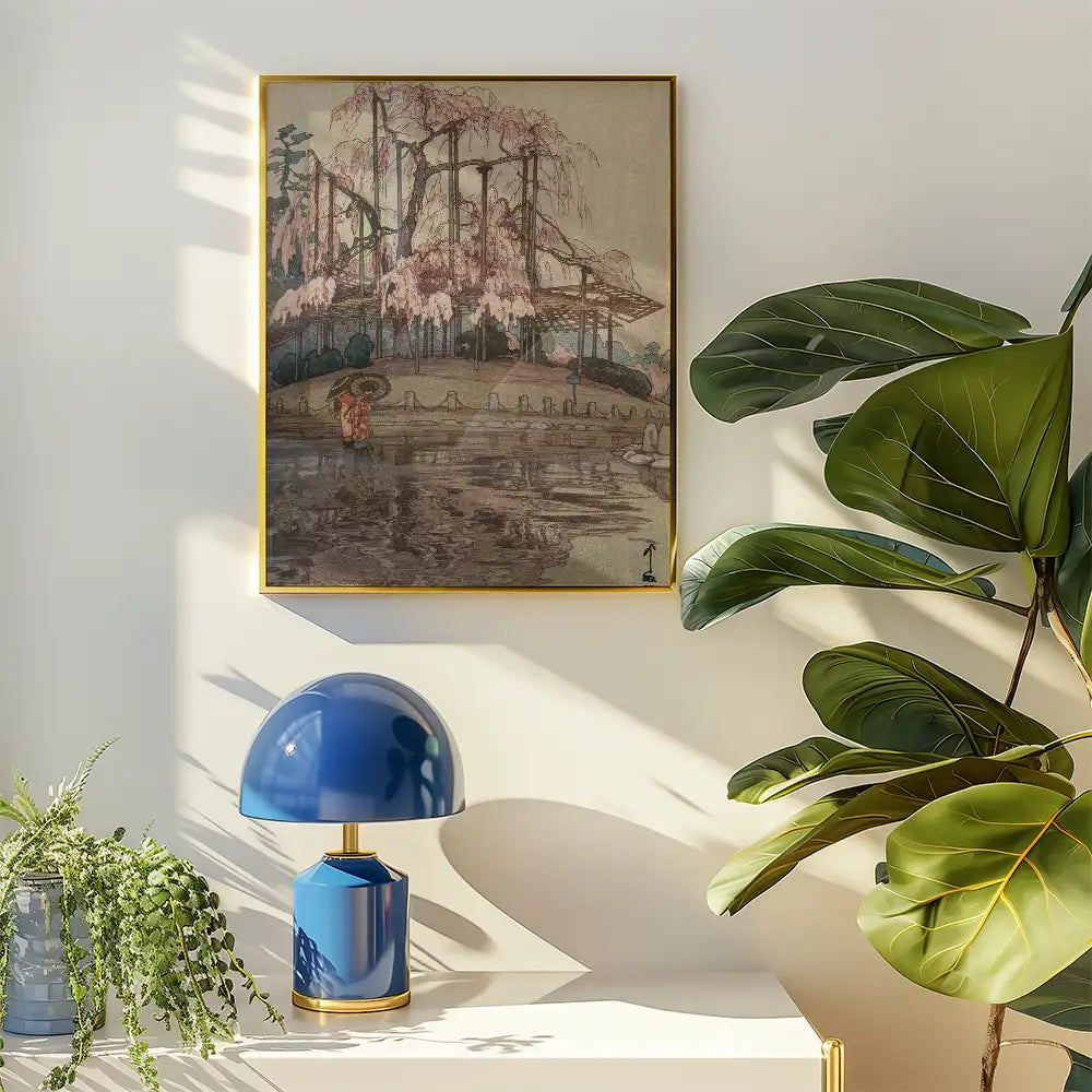 Painting of cherry blossoms by a tranquil pond, framed in gold, displayed on a white wall near a blue lamp and greenery.