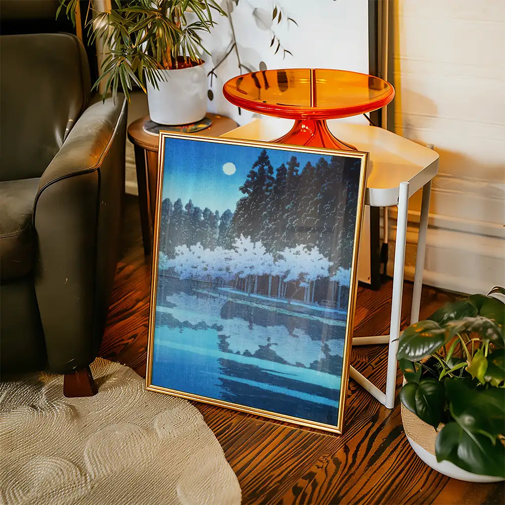 Framed landscape painting featuring a moonlit lake and trees, displayed in a cozy living room setting with a modern accent table and greenery.
