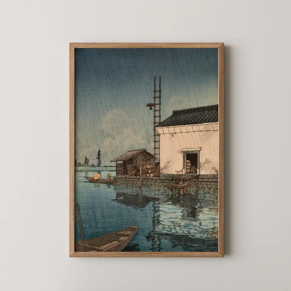 Framed painting of a serene waterside scene with the rain falling, featuring a rustic house, a boathouse, and a small boat on calm water reflecting the sky.