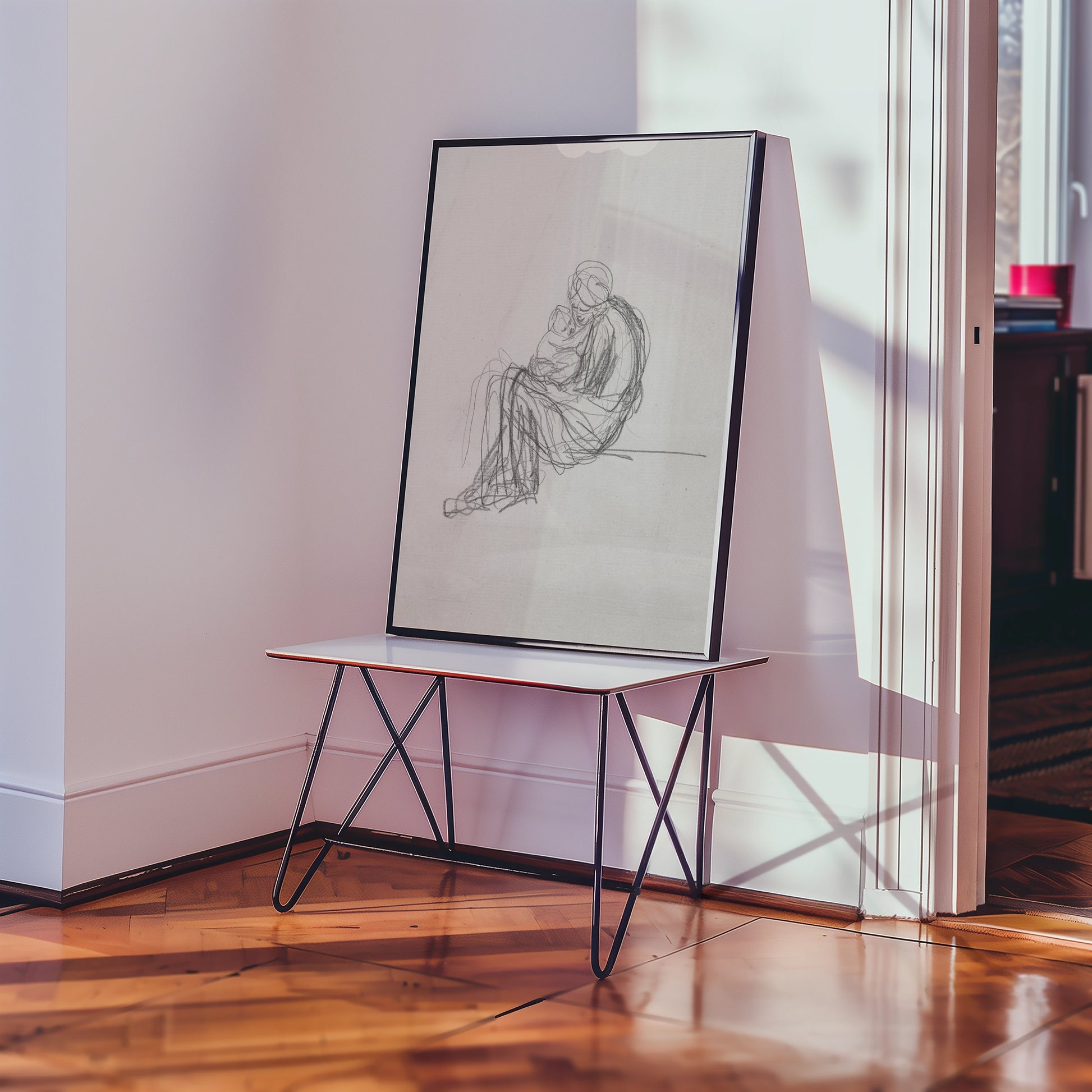 Framed black and white sketch painting of a seated figure with a child, placed on a minimalist table in a bright room.