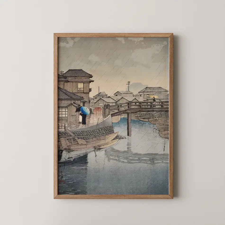 Framed painting of a rainy riverside scene featuring traditional Japanese houses, a wooden bridge, and a figure with an umbrella standing on a boat.