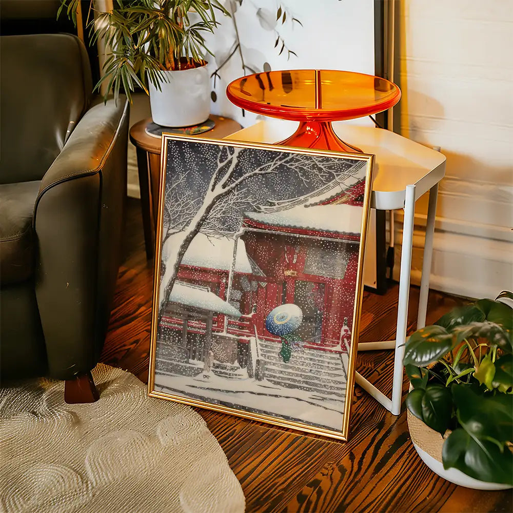 Framed painting of a snow-covered traditional building with a person holding an umbrella, placed beside a mid-century modern chair and a side table in a cozy living space.